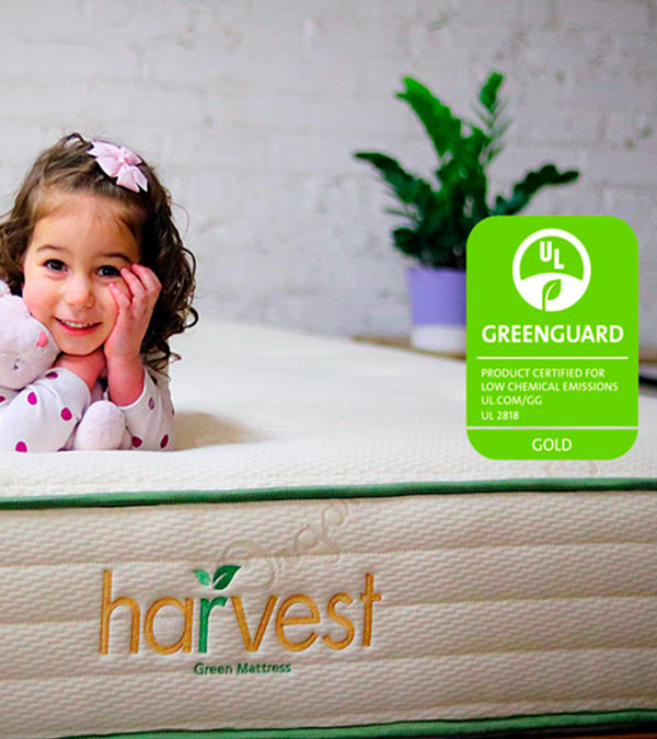 Little girl laying on our harvest green original mattress holding a teddy bear and smiling with our Greenguard Gold Certification logo